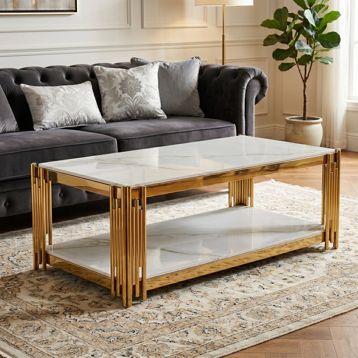 A modern rectangular coffee table with a white marble-patterned top and lower shelf, supported by a polished gold-toned metal frame, set in a styled living room next to a dark gray sofa. Belini Coffee table by House of Murphy