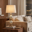 Switch A table lamp with a textured beige fabric drum shade, a hammered silver glass stem with two stacked oval bulbs, and a bronze-finished base on a white background. 2 image